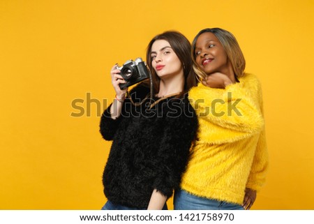 Two young women friends european and african american in black yellow clothes standing posing isolated on bright orange wall background, studio portrait. People lifestyle concept. Mock up copy space