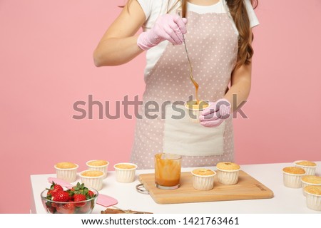 Close up cropped cute housewife chef cook confectioner or baker in white t-shirt cooking at table isolated on pink pastel background in studio. Cupcake making process. Mock up copy space food concept
