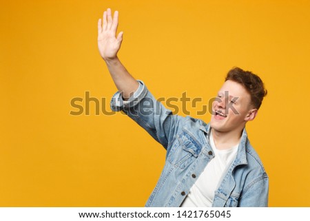 Pleasant young man in denim casual clothes waving and greeting with hand as notices someone isolated on yellow orange wall background. People sincere emotions, lifestyle concept. Mock up copy space