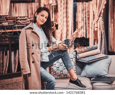 Stylish cheerful designer is sittingon the sofa at her cozy textile workplace surrounded by many different garments.