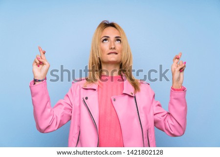 Young blonde woman with pink jacket over isolated blue background with fingers crossing and wishing the best