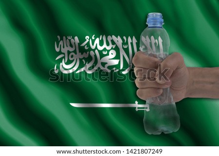 Concept of stopping plastic pollution. Ban disposable products. A hand squeezes a plastic bottle to protest the use of disposable plastic containers against the background of the flag of Saudi Arabia