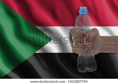 Concept of stopping plastic pollution. Ban disposable products. A hand squeezes a plastic bottle to protest the use of disposable plastic containers against the background of the flag of Sudan
