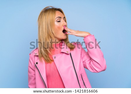 Young blonde woman with pink jacket over isolated blue background yawning and covering wide open mouth with hand