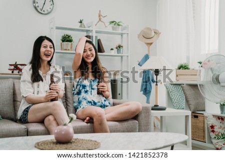 two young asian women relaxing near air cooler at home. beautiful girl friends holding cold ice beer sitting on couch sofa enjoy watching movie comedy cartoons on television looking tv screen indoor