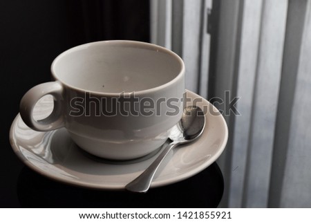 White coffee mug placed​ on dark brown old wooden table​ with​ Old brown chair​ and​ window, curtains​ with​ sunlight​ on background.