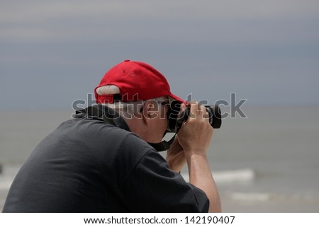 grey haired man in red hat and black shirt taking pictures of beach