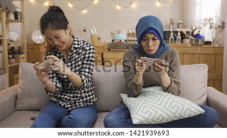 joyous asian female friends playing together video games on mobile phones with kitchen background. two chinese and malay roommates sitting on couch using cellphone having fun relaxing at home