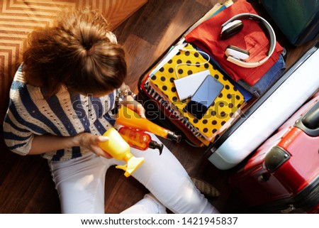 Upper view of modern woman in white pants and striped blouse in the modern house in sunny summer day packing summer cosmetics in open travel suitcase.