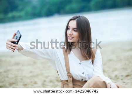 Happy smiling hipster girl making self portrait with mobile phone camera while sitting in city park, cheerful woman posing while photographing herself for social network picture.