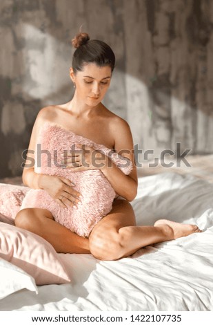 Beautiful pregnant girl without clothes is sitting on the bed in the early morning and covers her tummy with a pillow.