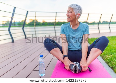 Relaxed athletic mature woman sitting on fitness mat outdoors. Senior Woman Resting After Exercises. Woman on a yoga mat to relax outdoor. Senior lady prefers healthy lifestyle Royalty-Free Stock Photo #1422159878