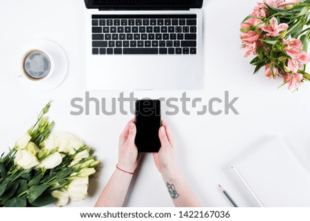 cropped view of woman holding  smartphone with blank screen near laptop, cup of coffee and bouquets on white
