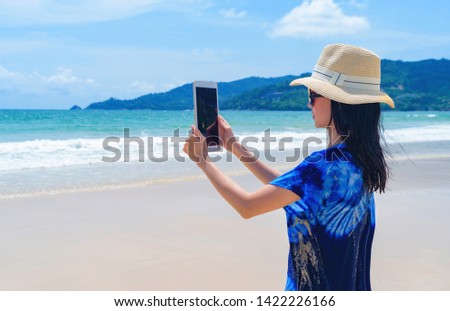 Happy Asian woman using mobile phone to take a photo by camera on social media at beach during travel holidays vacation outdoors at ocean or nature sea at noon, Phuket, Thailand