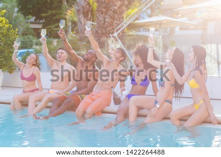 Multiethnic group of young people toasting with glasses of champagne sitting on the edge of the swimming pool