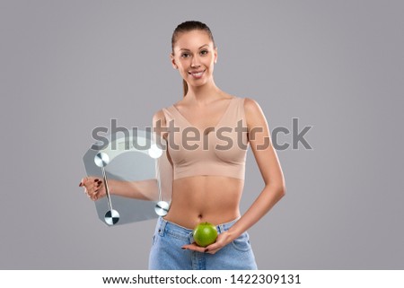 Dieting young female smiling and looking at camera while demonstrating modern scales and healthy apple against gray background
