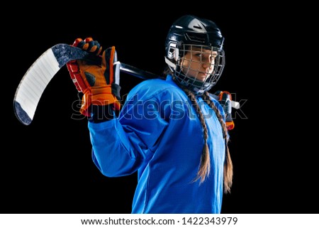 Young female hockey player isolated on black background. Sportswoman wearing equipment and helmet standing with the stick. Concept of sport, healthy lifestyle, motion, movement, action.