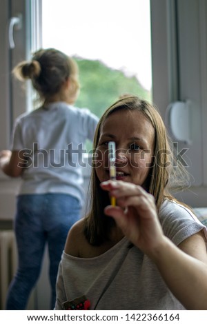 Mother is holding syringe while her daughter is looking through window. Close-up of mother holding medication. Cute girl turned back to her mother, looking out the window, while mother takes medicine.