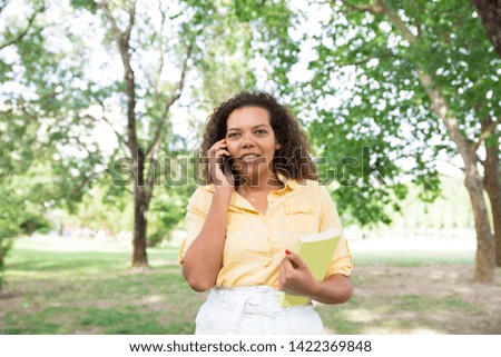 Positive woman talking on phone and holding book in city park. Young woman standing with blurred trees and lawn in background. Summer and information concept. Front view.