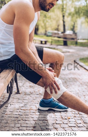 cropped shot of sportsman sitting on bench and touching elastic bandage on injured leg