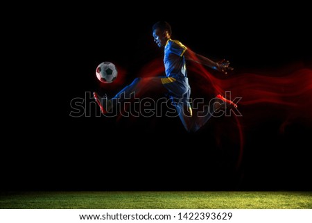 Young african-american male football or soccer player in sportwear and boots kicking ball for the goal in mixed light on dark background. Concept of healthy lifestyle, professional sport, hobby.