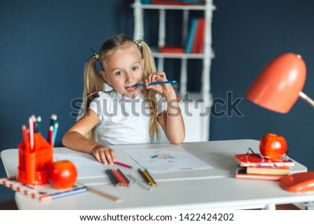 Funny little girl with blond hair sitting at white table and holding purple pencil in her mouth