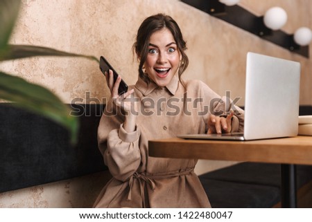 Image of a shocked young beautiful woman indoors in cafe using laptop computer talking by mobile phone.