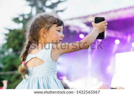 A little girl with a phone in hand is having fun at a musical concert. A child takes on the camera a street festival of popular music. Daughter sits on dad's neck