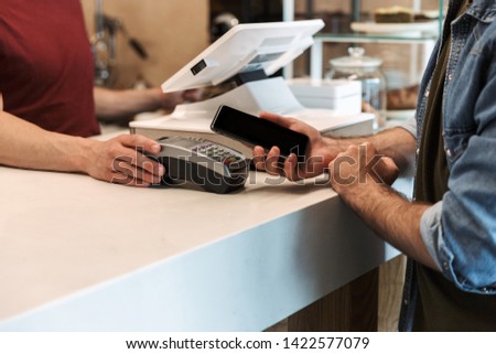 Cropped photo of young man wearing denim shirt paying debit card in cafe while waiter holding payment terminal
