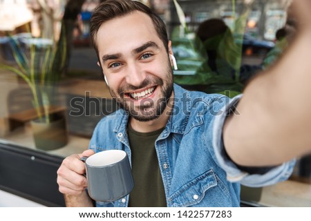 Photo of handsome young man wearing earpods smiling and drinking cup of coffee while taking selfie on smartphone in city cafe outdoors