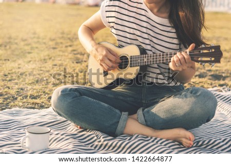 Brunette hipster girl in a striped T-shirt and on a hot day on the beach in soft sunset lighting plays a small guitar ukulele. Relaxed atmosphere, lifestyle image, copy space for text. 