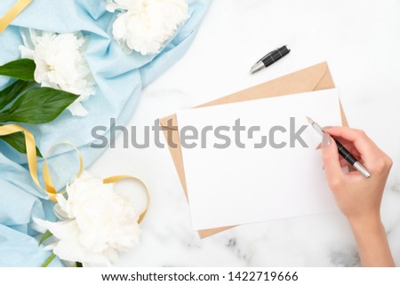 Top view woman hand writing wedding invitation card or love letter. Minimal flat lay composition with peony flowers, stationery stuff and feminine accessories. Writing letter concept