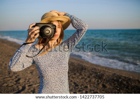 Young attractive girl with long blond hair, wearing a hat, takes pictures at the beach. Holding photocamera in one hand. Blue waves on the background.