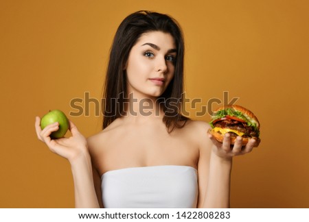 Young woman making decision between healthy food green apple and fast food burger. Fitness, diet and food concept - doubting woman with fruits and hamburger on yellow background