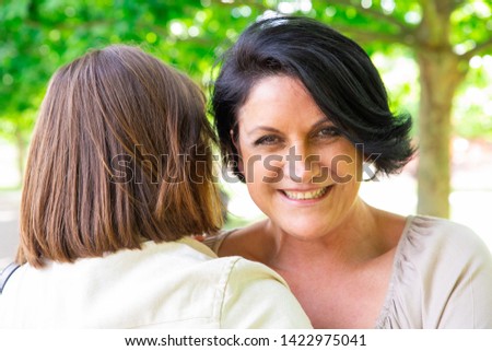 Happy middle-aged woman embracing daughter in park. Middle-aged lady embracing her daughter who is standing back to camera with green trees in background. Family relationship concept.