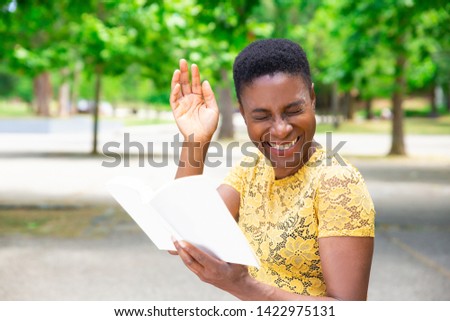 Laughing African-American woman reading joke in book. Happy beautiful woman spending time in park with book of funny stories. Literature concept