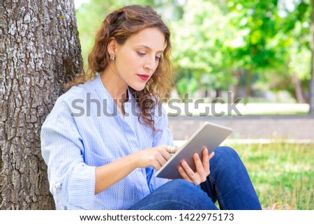 Curious young woman reading online article on tablet. Serious wavy-haired girl in casual shirt sitting on grass and leaning on tree in park. Surfing net concept