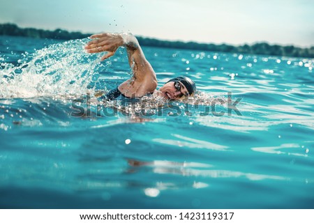 Professional triathlete swimming in river's open water. Man wearing swim equipment practicing triathlon on the beach in summer's day. Concept of healthy lifestyle, sport, action, motion and movement. Royalty-Free Stock Photo #1423119317