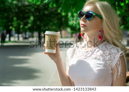 Beautiful blonde girl in the city is drinking coffee. Street / Urban photo session. Gray cup with a white lid and a place for the logo