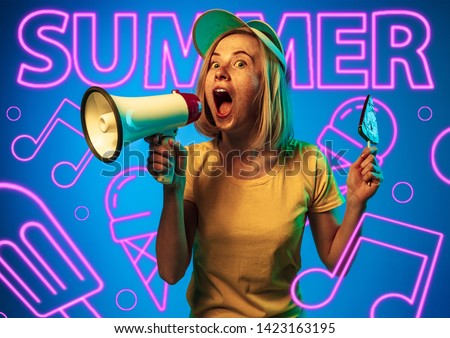 Female model in trendy neon light on blue background. Modern design. Beautiful young woman posing in cap, screaming and holding a candy. Concept of facial expression, summer, vacation, music, open-air