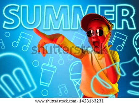 Female model in trendy neon light on blue background. Modern design. Beautiful little girl posing in cap and sunglasses, looks astonished. Concept of facial expression, summer, vacation, music.