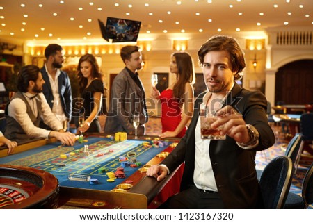 A man in a suit holds a glass with alcohol in his hand against the background of a roulette game in a casino.
