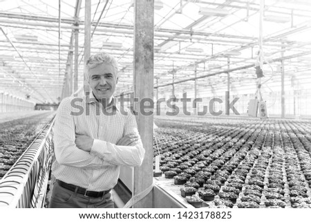 Black and White photo of Portrait of confident mature owner standing with arms crossed at greenhouse