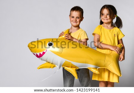 Positive young children hold a balloon in the shape of a yellow shark fish, celebrate the holiday, smiling widely, stand on a light background, are in a good mood.