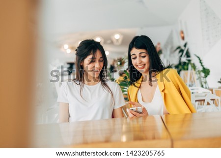 Cheerful nice young businesswomen sitting in coworking together. Look at smartphone and working. Happy positive person in white room