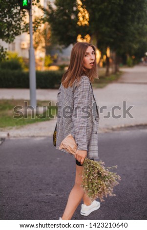 Attractive beauty woman cross the road while wearing jacket in square. Photo from back and girl is turn around. Business woman at evening and go to home after job with baguette and bouquet of flowers.
