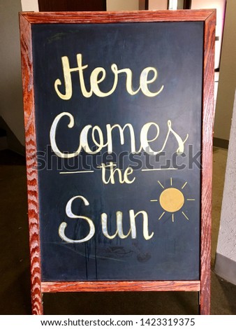 "Here comes the sun" chalk board bulletin board outside of a cafe restaurant in handwritten letters with a sun graphic and a stained wooden frame.