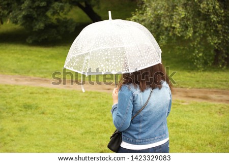 A girl in a denim blue jacket with a white transparent umbrella walks in the rain among the green. Romantic mood. Melancholic wet weather. Walk in rainy weather with a trendy gadget. Autumn-spring