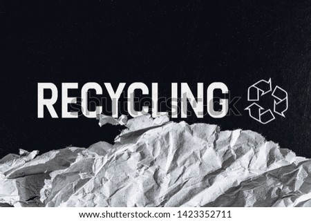 Black textured carton background with printed white recycling logo sign and piece of recycled crumpled paper. Space for text. Minimalistic style template. Bold text RECYCLING. Horizontal orientation