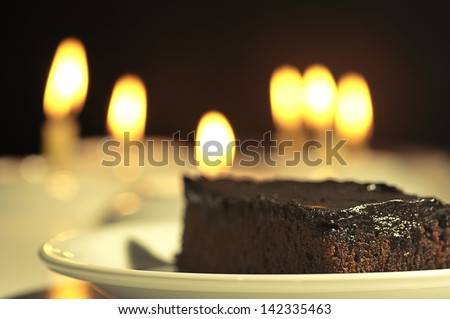 happy birthday  -  a brown / black cake slice. on a white plate, and candles lights in the background.